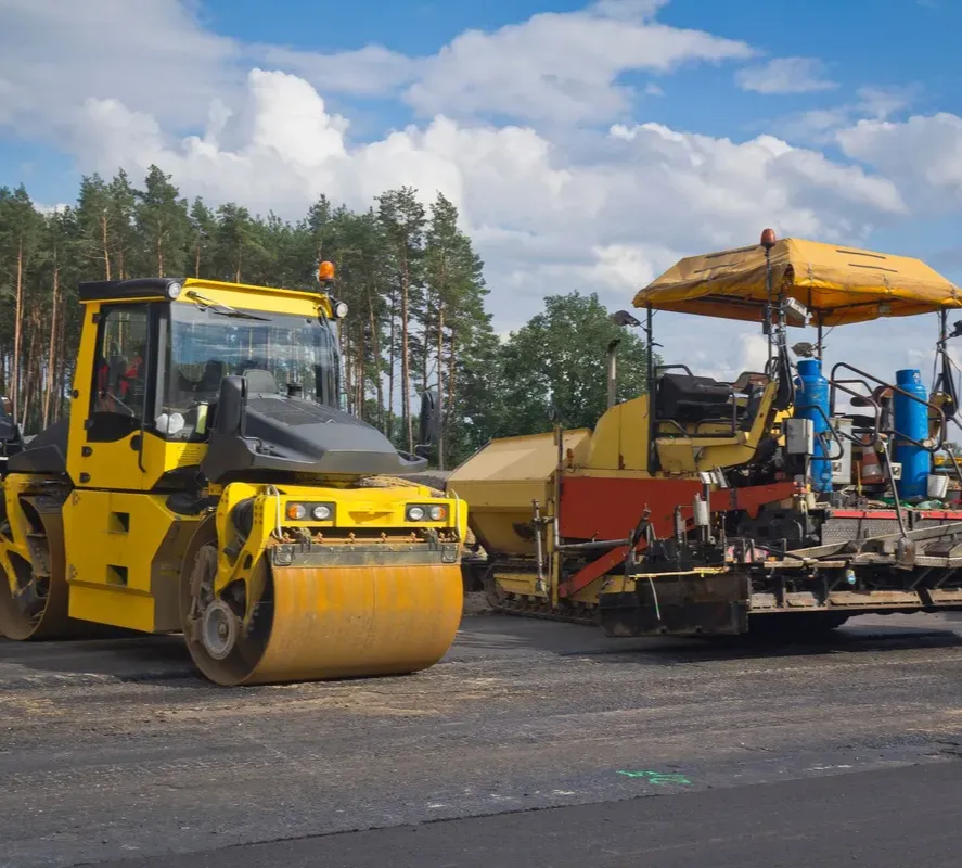 about-1 Yellow road roller and asphalt paver working on a road construction site under a partly cloudy sky.