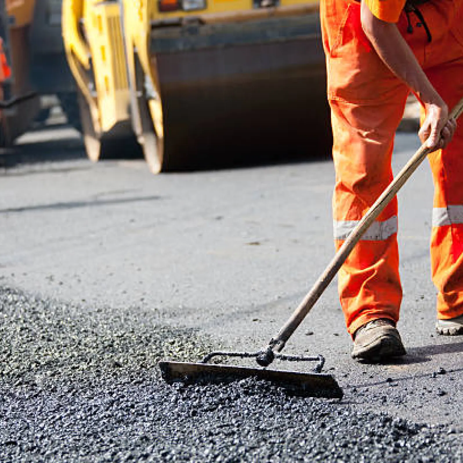 asphalt-inst-1 Worker in orange gear smooths asphalt with a rake on a road construction site, steamroller in background.