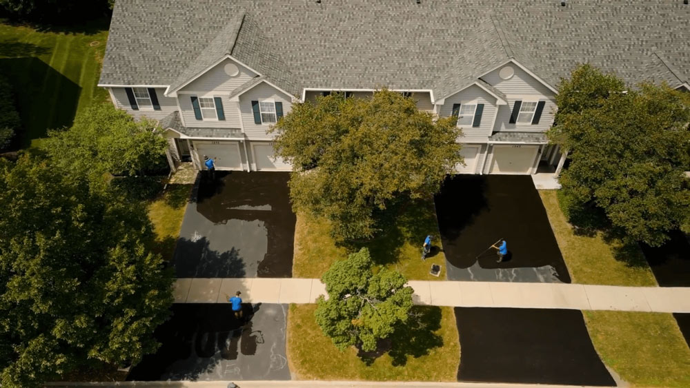 Aerial view of workers sealing driveways in front of a row of suburban townhouses with trees and grass.