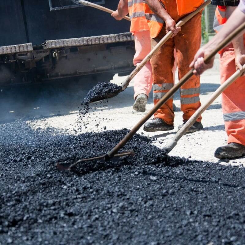 Workers in orange uniforms spreading hot asphalt with shovels on a road construction site.
