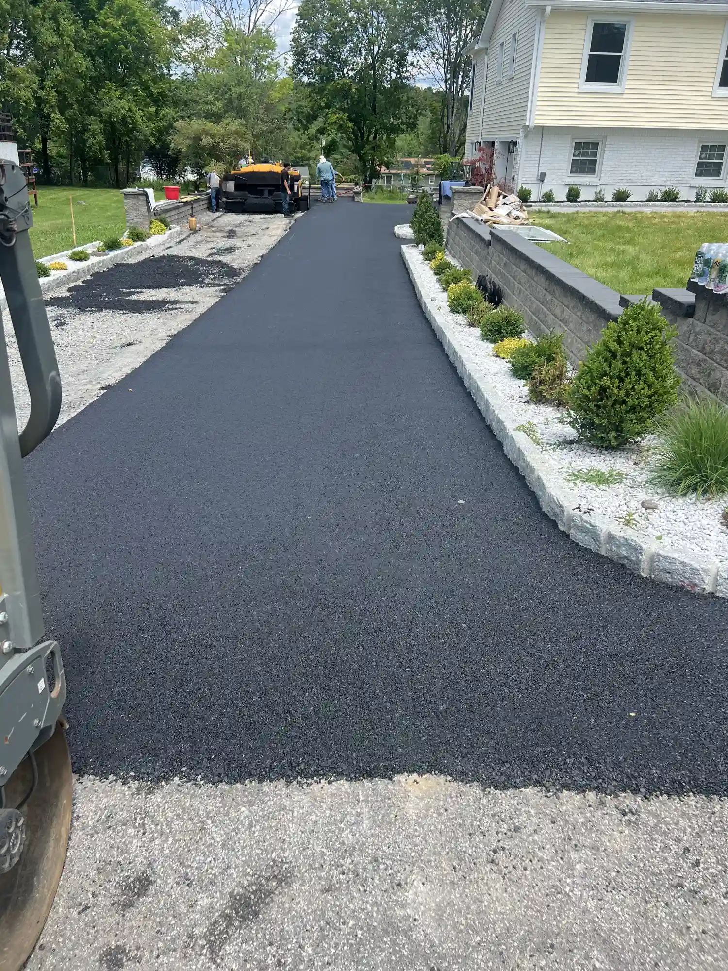 Fresh black asphalt paving on a residential driveway with landscaping and workers in the background.