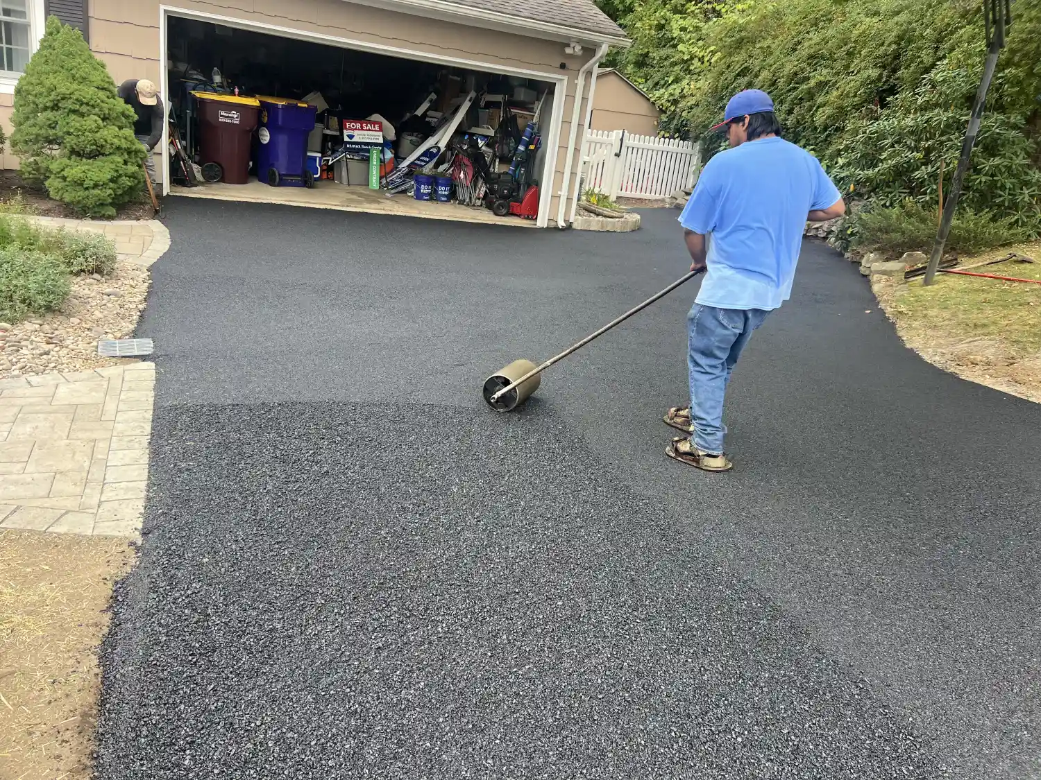 asphalt paving contractor: Person in blue shirt spreads asphalt on a driveway with a roller in front of an open garage.