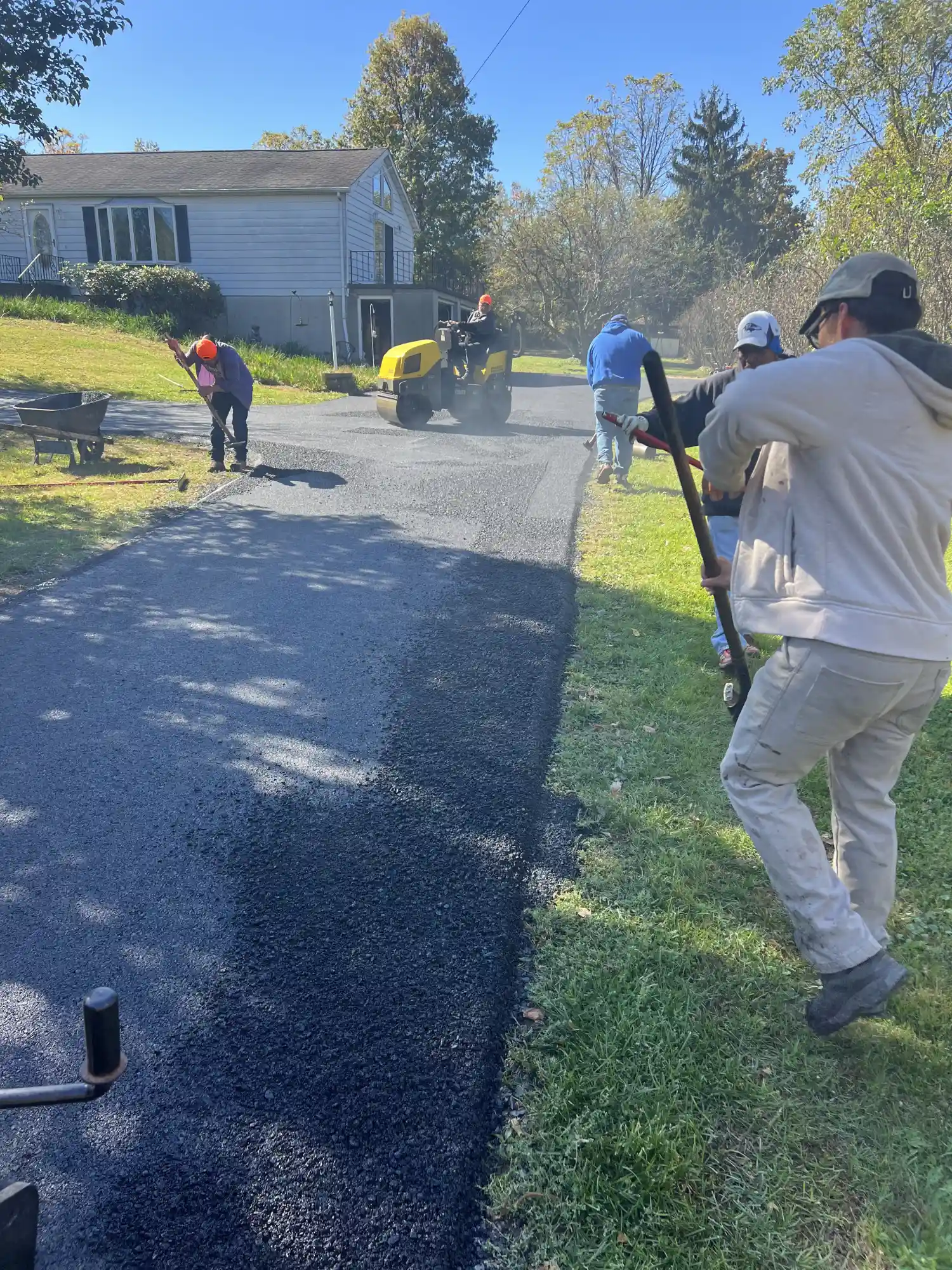 Workers paving a driveway with asphalt near a house on a sunny day, using tools and a steamroller.