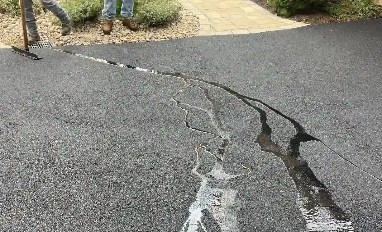 Water flows through cracks in a newly paved asphalt driveway; a person stands nearby with tools.