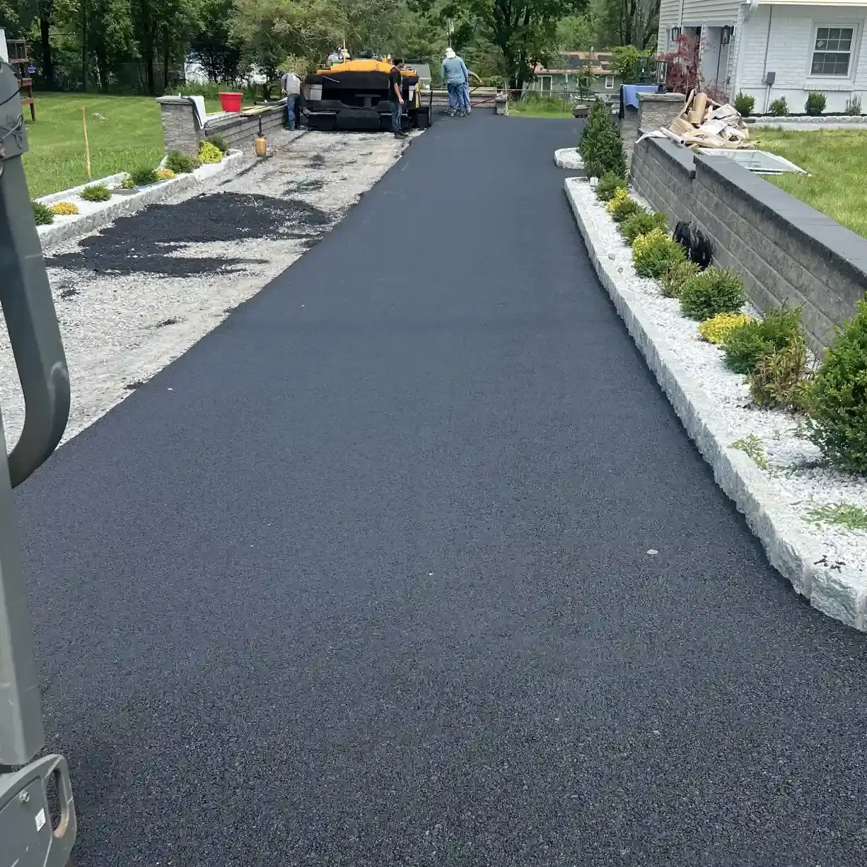 Freshly paved asphalt driveway with workers and paving equipment near a white house and landscaped yard.