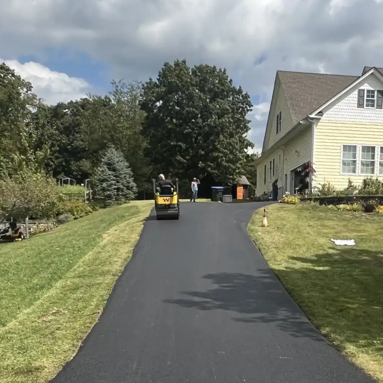 A worker drives a roller on a freshly paved driveway beside a yellow house on a sunny day.