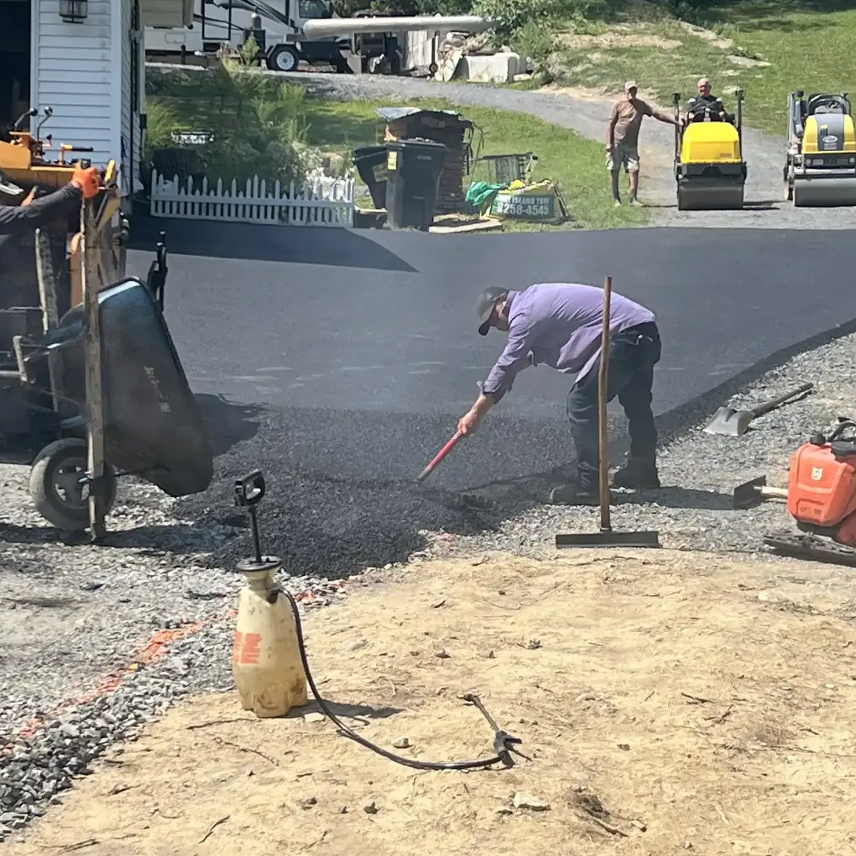 asphalt installation: A worker spreads asphalt on a driveway while others and construction equipment are visible in the background.
