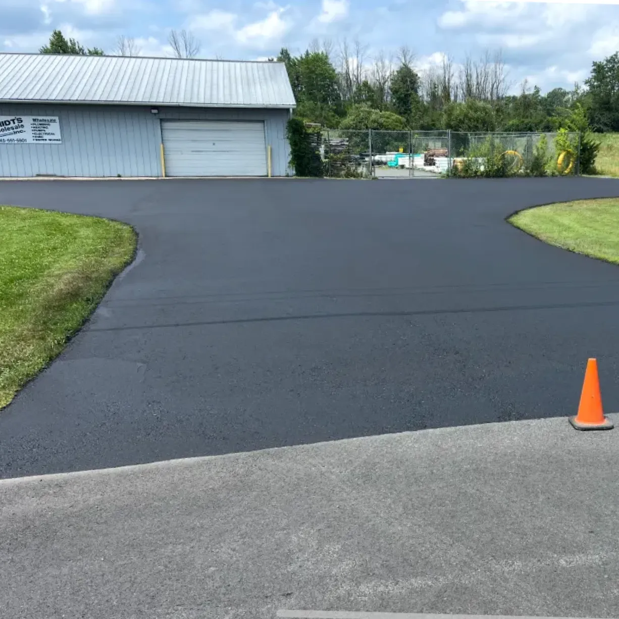 Freshly paved asphalt driveway leading to a metal building, with a single orange cone in the foreground.