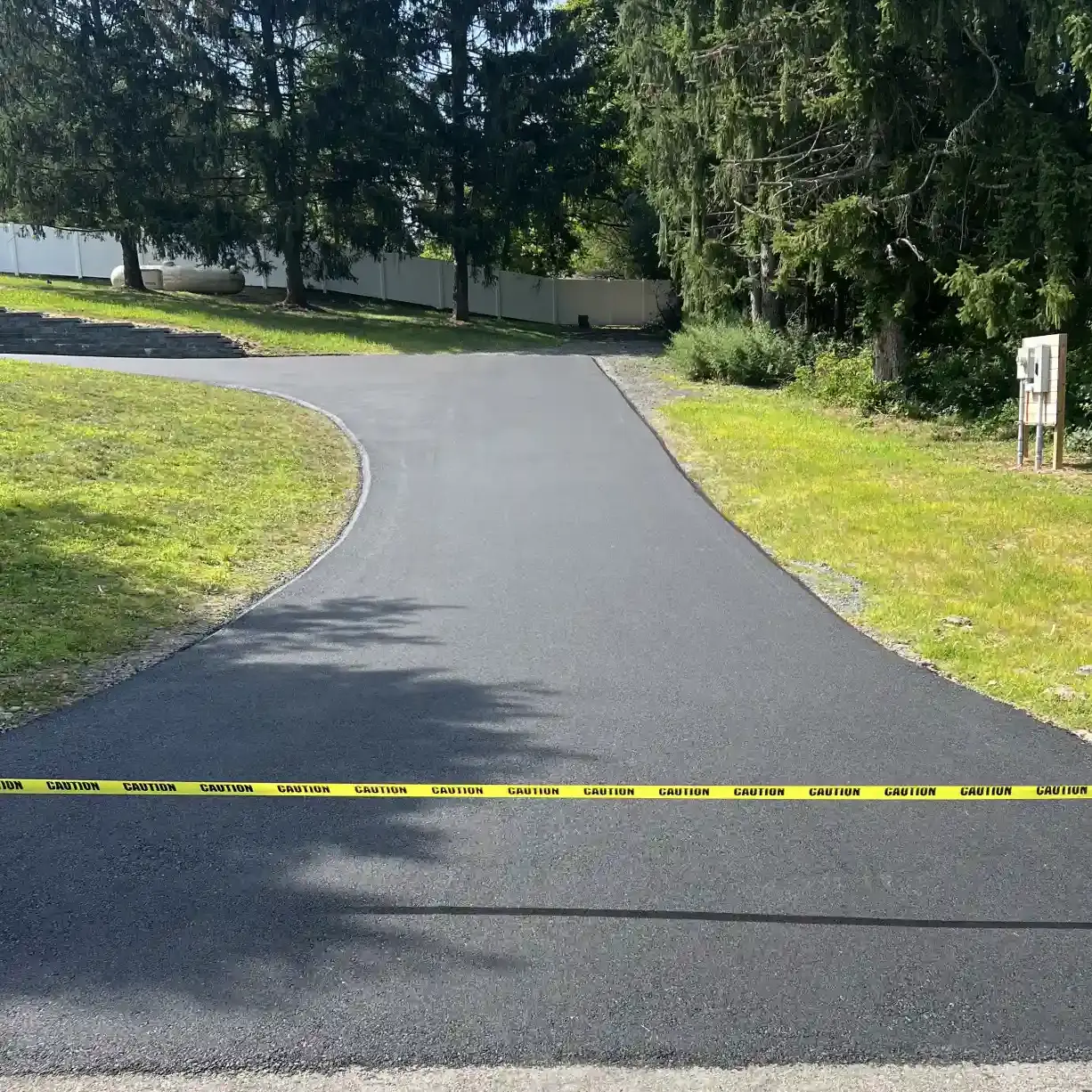 Newly paved driveway blocked by yellow caution tape, surrounded by grass and trees.